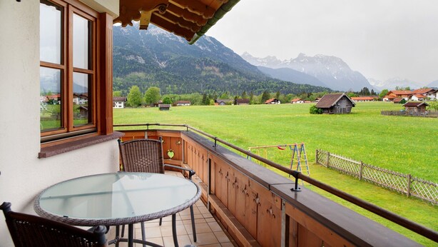 A balcony with a glass table and chairs, a wooden railing, and a view of a green field and mountains. - Apartment 'Landhaus Bergkulisse' with Mountain View, Shared Terrace and Wi-Fi (Wallgau)