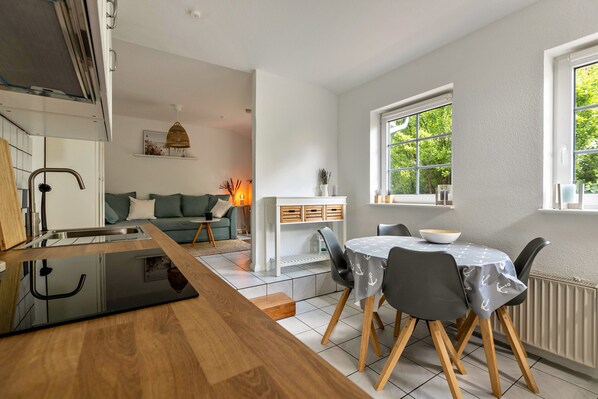 A modern kitchen with a wooden countertop and integrated appliances, a dining area with a round table and chairs, and a view of greenery through a large window.