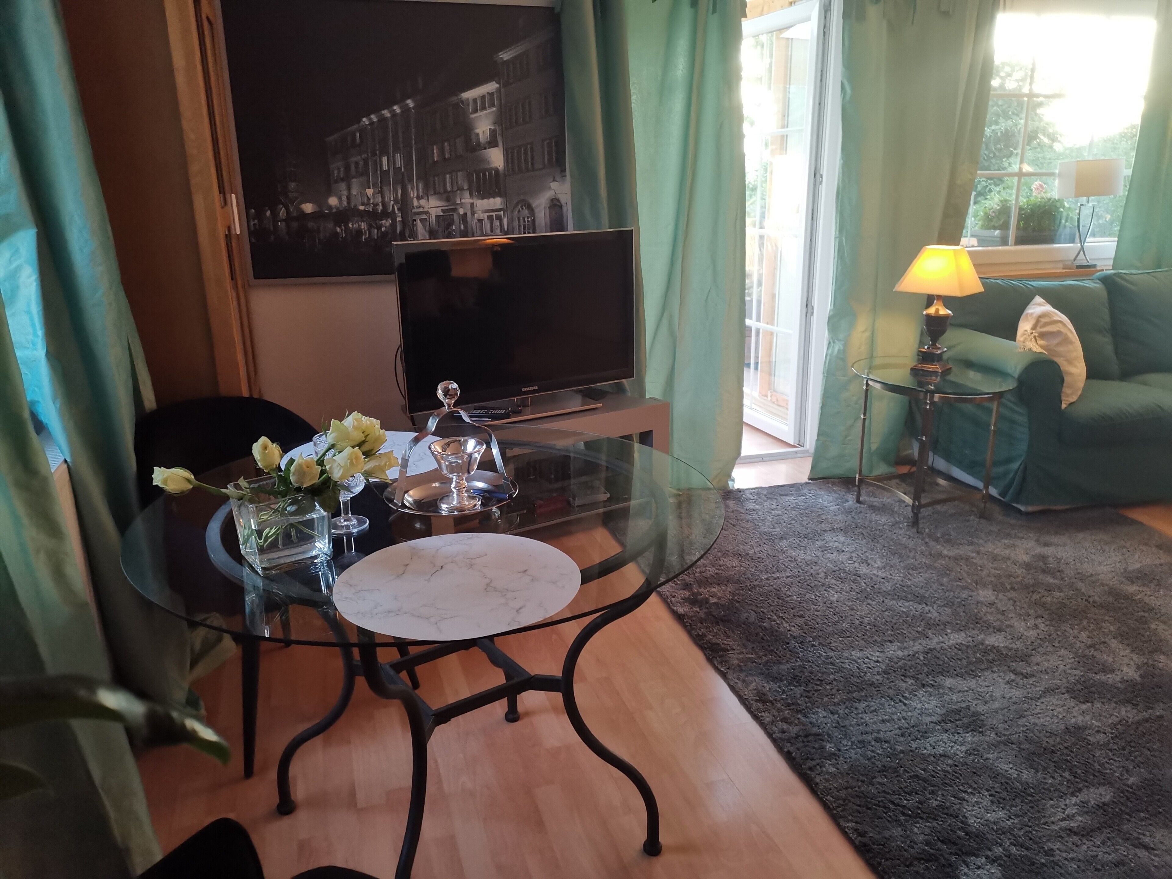 A room with a glass table, a flat-screen TV, a sofa, and a lamp.