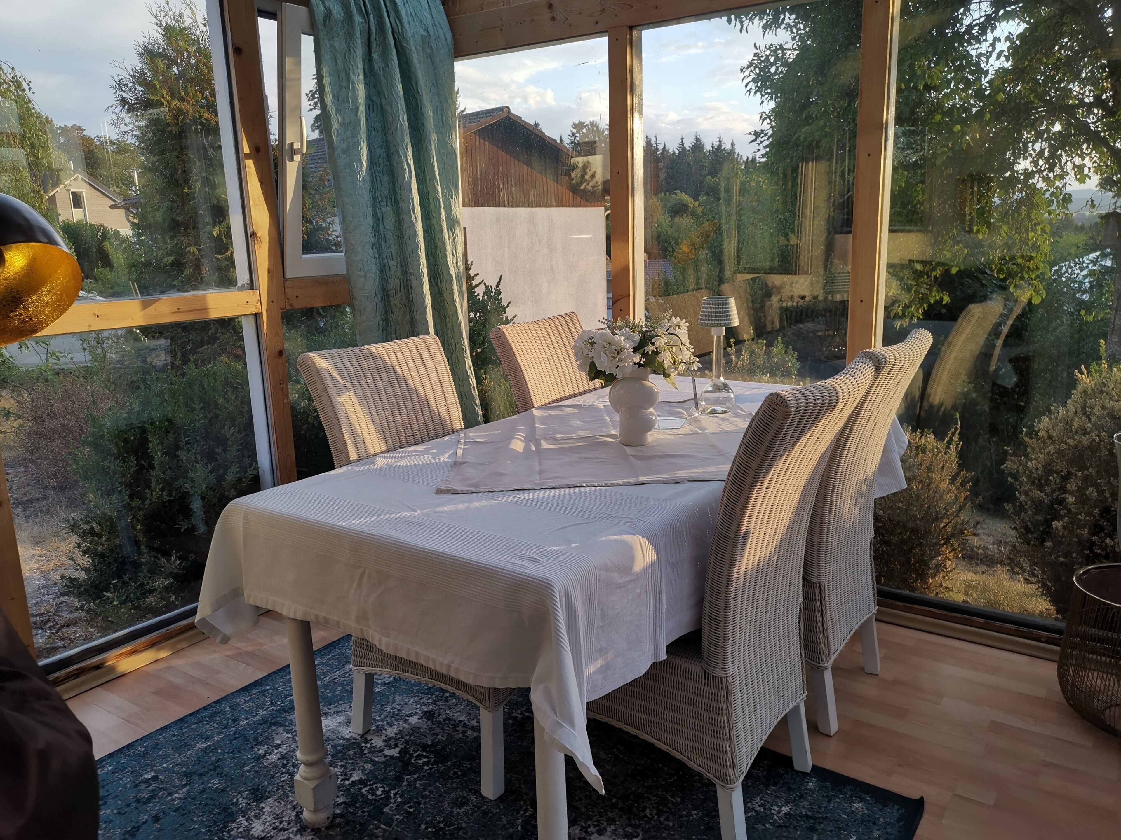 Un comedor con una mesa dispuesta para dos personas, sillas de mimbre, un jarrón con flores y una lámpara.