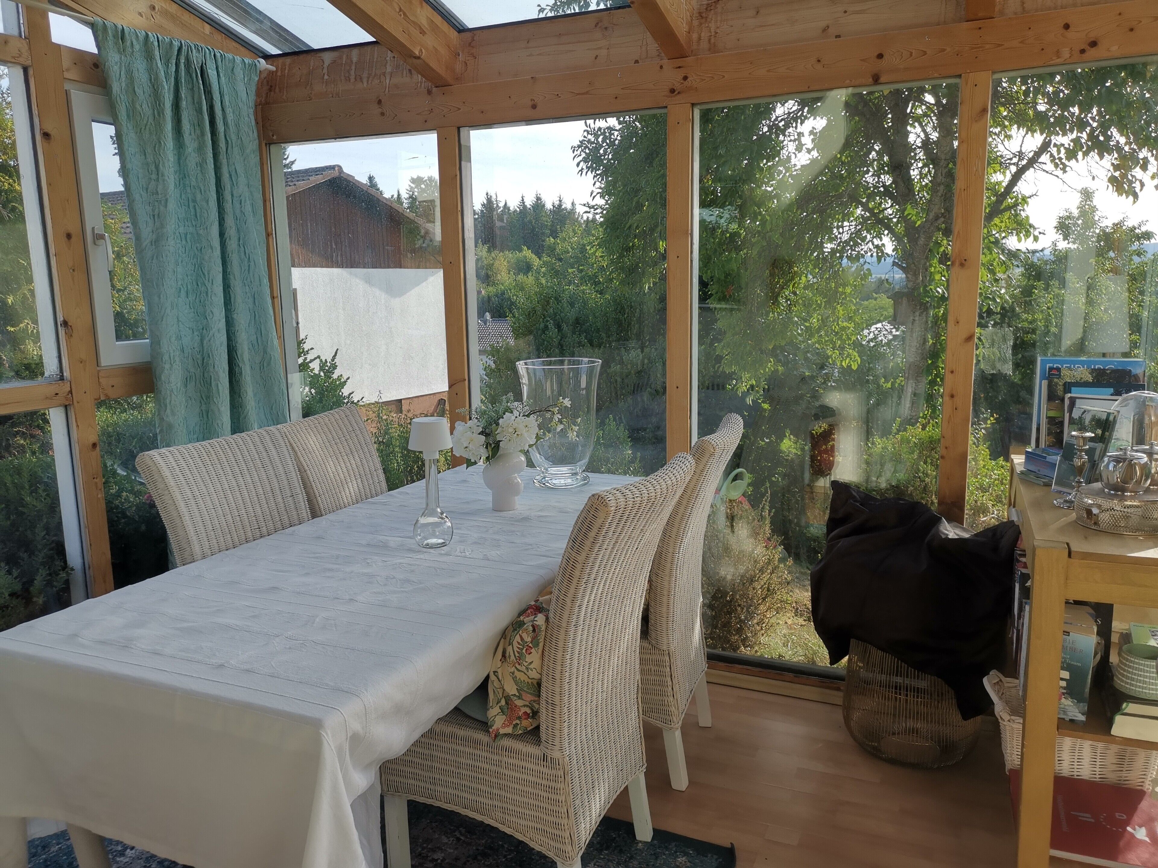 Une véranda avec une table dressée pour deux personnes, des chaises en osier, un vase rempli de fleurs et une vue sur la verdure environnante.