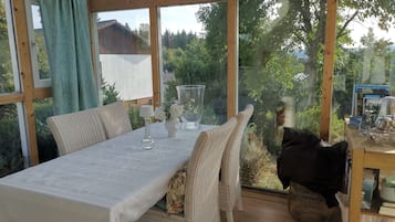 A conservatory with a table set for two, wicker chairs, a vase with flowers, and a view of greenery outside.