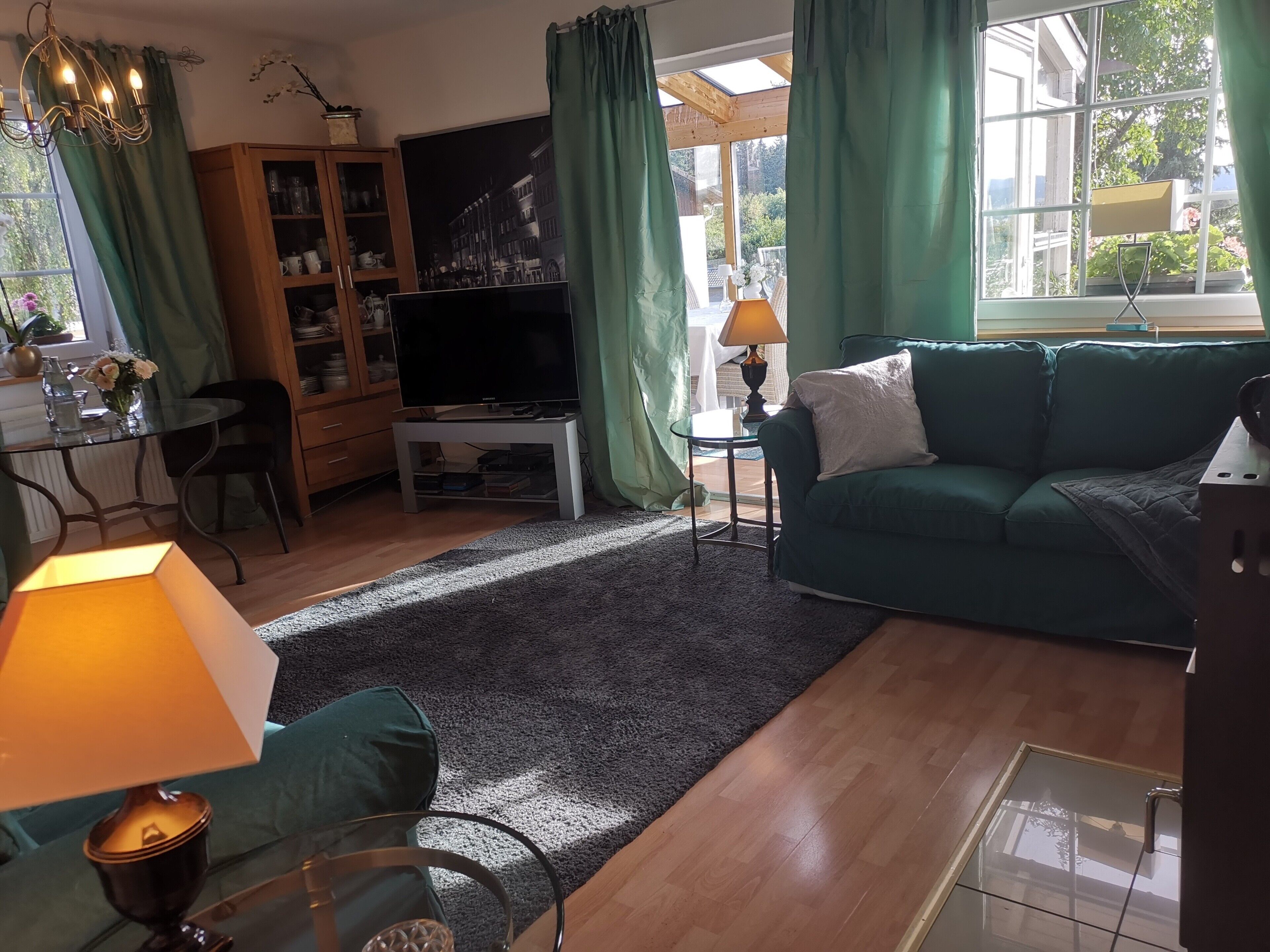 Een woonkamer met een groene bank, een televisie op een dressoir, een glazen tafel en een kast met glazen deuren.