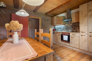 Una mesa de comedor de madera con sillas, un florero con flores secas, una cocina con armarios de madera y salpicadero de piedra, y una lámpara colgante.