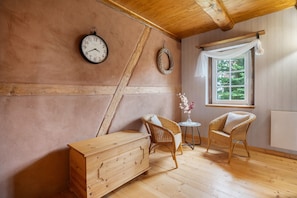 Una habitación con techo de madera, un aparador de madera, dos sillas de mimbre, una mesita con un florero y una ventana con vistas a la vegetación.