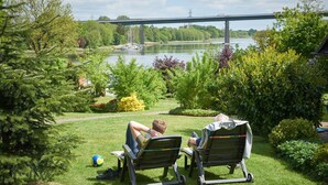 Two people relaxing on lawn chairs by a river with a bridge in the background. - Holiday Home 'Eiderblick' with Private Terrace, Private Garden and Wi-Fi (Borgstedt)