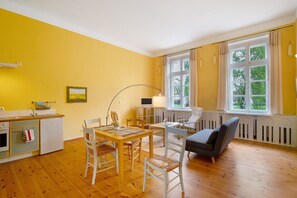 Ein Zimmer mit Holzboden, einem Esstisch mit Stühlen, einer Couch, einer kleinen Küchenzeile mit Spüle und Kühlschrank sowie einem Fenster mit Vorhängen.