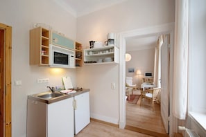 A compact kitchen with a microwave, stove, and shelves; a dining area with a table and chairs; and a view into a living space with a TV and seating. - Studio Apartment 'Ferienwohnung 3' with Shared Terrace, Shared Garden (Krakow am See)