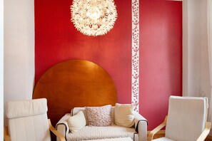 Une pièce avec une grande tête de lit en bois, un canapé blanc orné de coussins à motifs et deux fauteuils blancs.