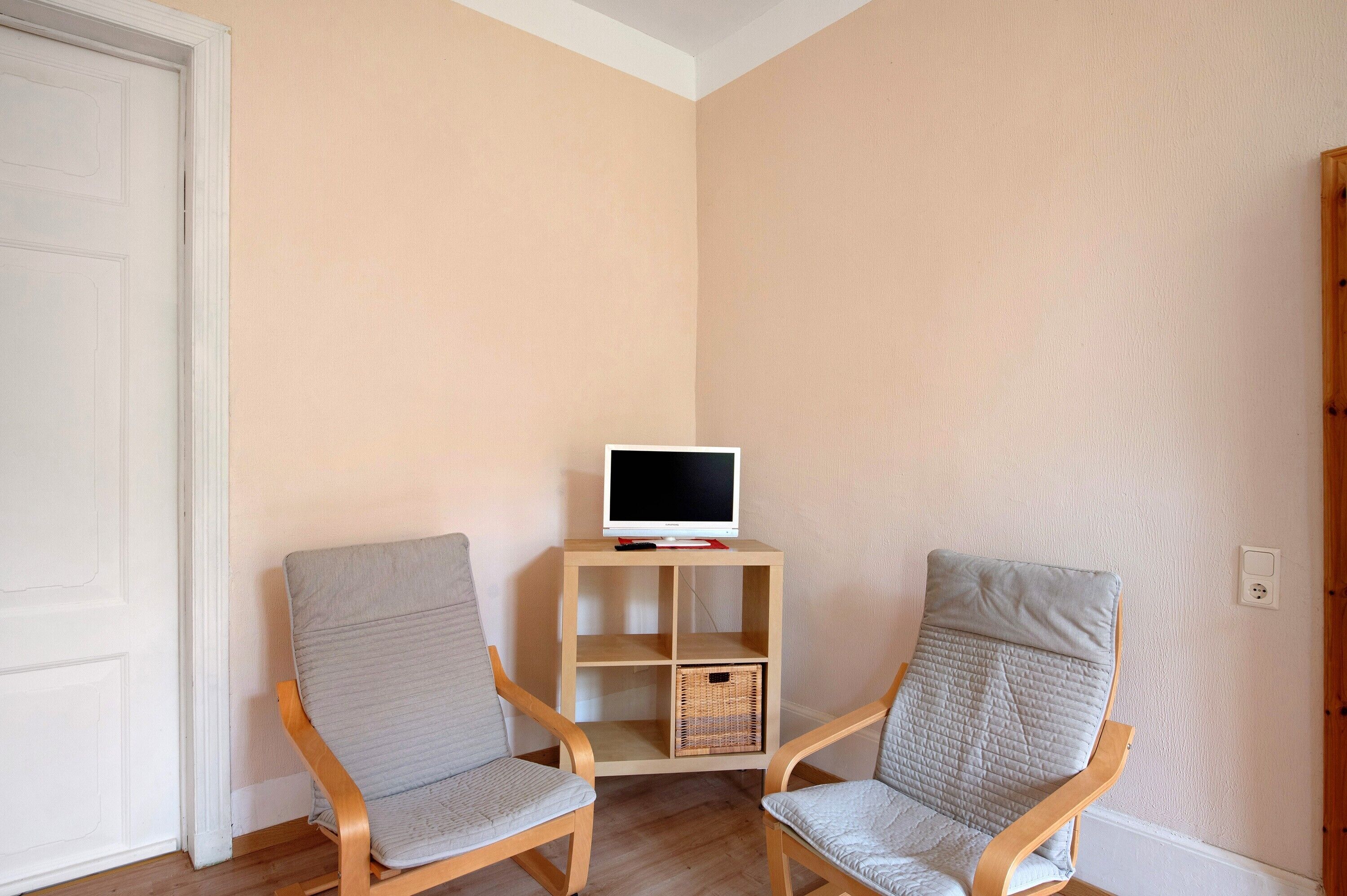 Ein Zimmer mit zwei Holzstühlen, einer kleinen Holzablage mit einem Fernseher und einer weißen Tür.