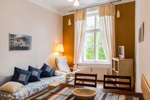Une pièce comprenant un canapé, un fauteuil, une table, une lampe, un téléviseur et une fenêtre avec des rideaux.
