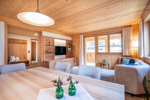 A wooden interior with a dining table, sofas, a TV, and windows.