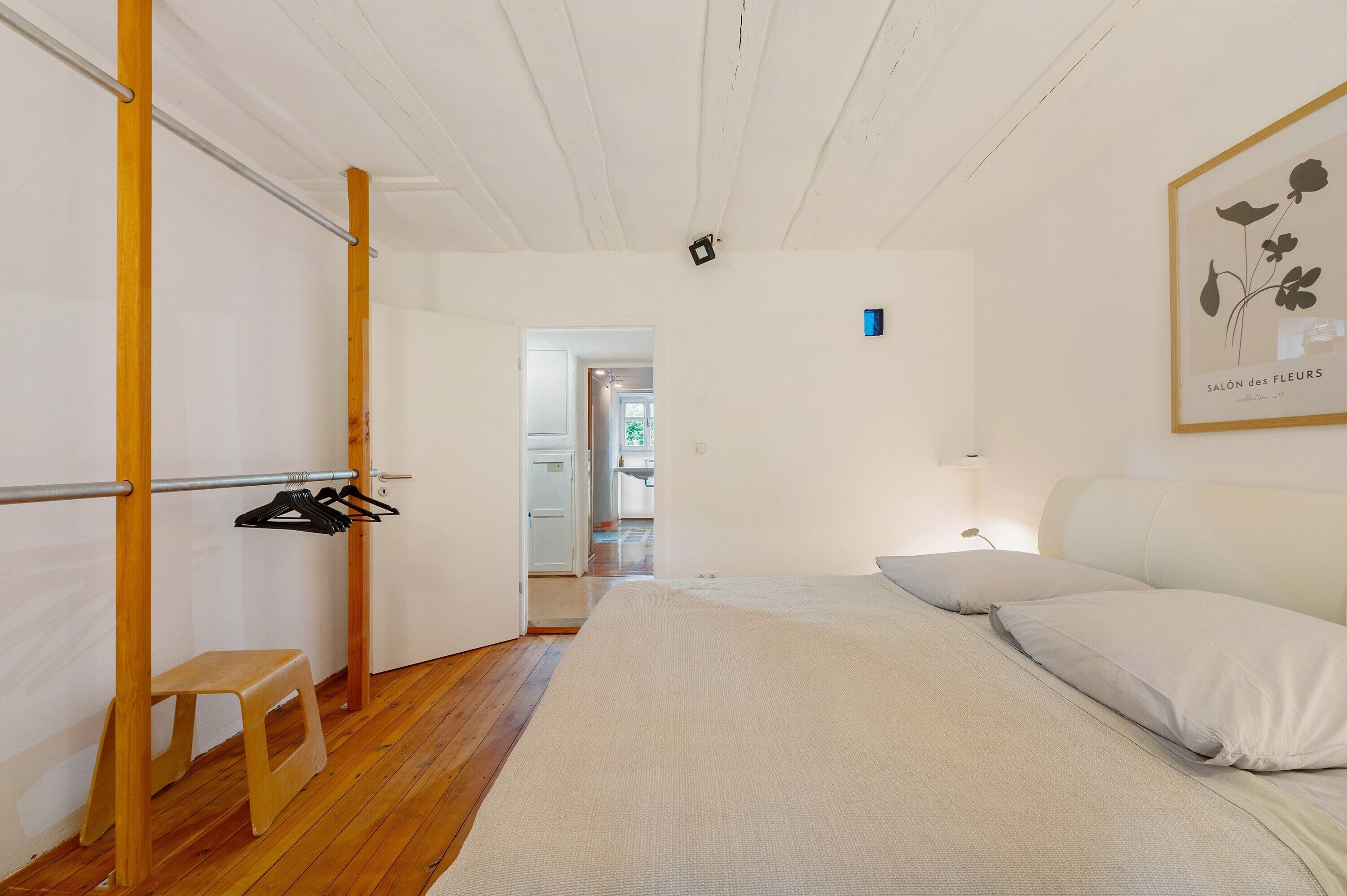 Une chambre à coucher comprenant un lit, un tabouret en bois, un portant à vêtements et une porte donnant sur une autre pièce.
