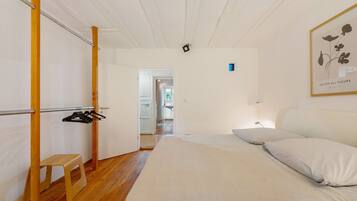 Une chambre à coucher comprenant un lit, un tabouret en bois, un portant à vêtements et une porte donnant sur une autre pièce.