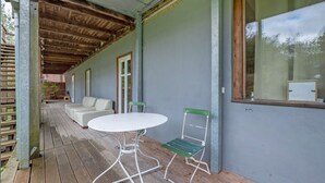 Een overdekt buitengebied met een witte tafel en twee stoelen, een houten bank en een raam met uitzicht op bomen.