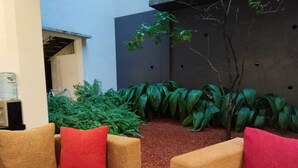 Uma área externa moderna com assentos, incluindo dois sofás bege e almofadas vermelhas, rodeada por plantas verdes exuberantes e uma árvore.