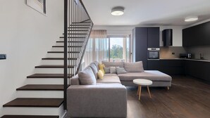 Un salon moderne avec un canapé d’angle gris, une petite table ronde et un escalier aux marches en bois foncé et aux rampes noires.