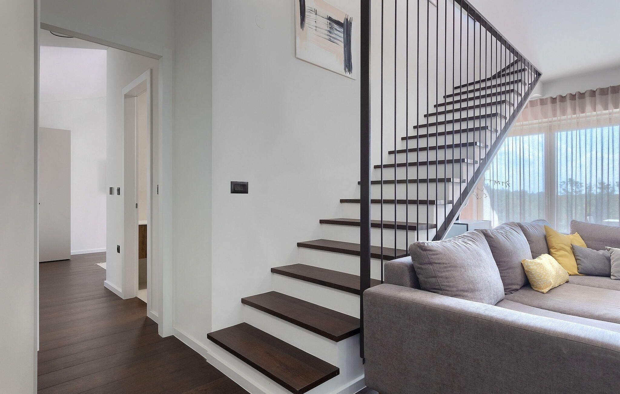 Un intérieur moderne avec un escalier, un canapé gris agrémenté de coussins et une fenêtre avec des rideaux.