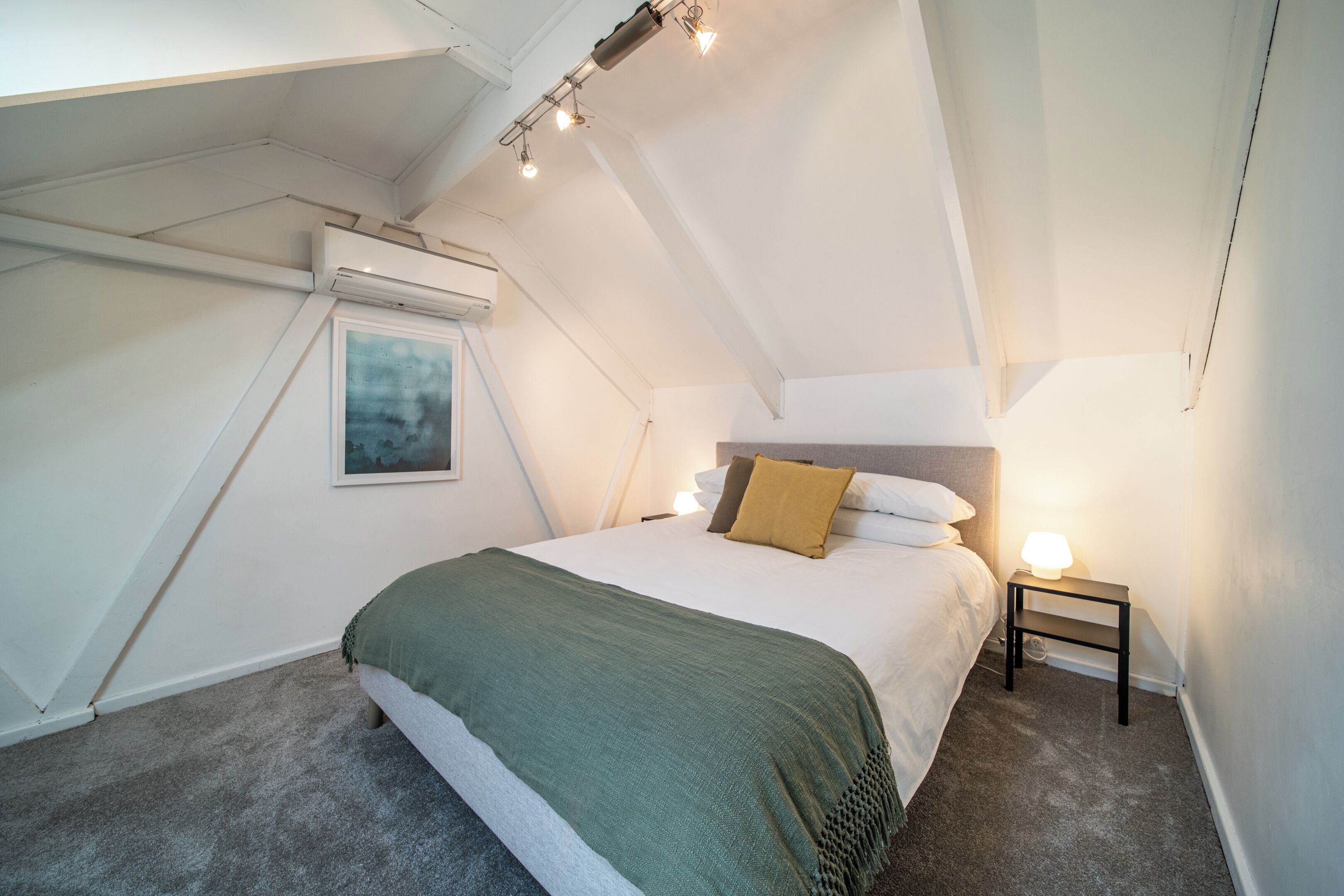 A bedroom with a bed, a nightstand, a lamp, a painting, and an air conditioning unit.