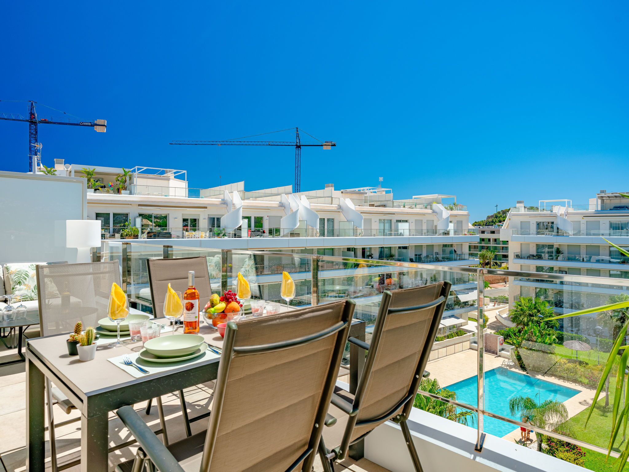 A rooftop dining area with a table set for two, overlooking a swimming pool and modern buildings.