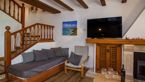En hyggelig stue med en trætrappe, en sofa med puder, en lænestol, et fladskærms-tv, en pejs og et bord med vinglas.
