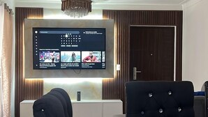 Una sala de estar moderna con un televisor de pantalla plana grande montado en la pared, un sofá capitonado, una mesa de centro de vidrio y una lámpara de techo.