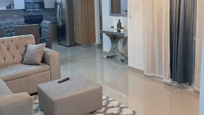 Una sala de estar moderna con un sofá modular, un pequeño reposapiés, una alfombra mullida, una araña y una cocina al fondo.