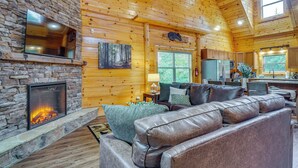 A log cabin interior with a stone fireplace, leather sofa, and a kitchen area. - 'Shady Pines' Mountain Retreat w/ Hot Tub! (Sevierville)