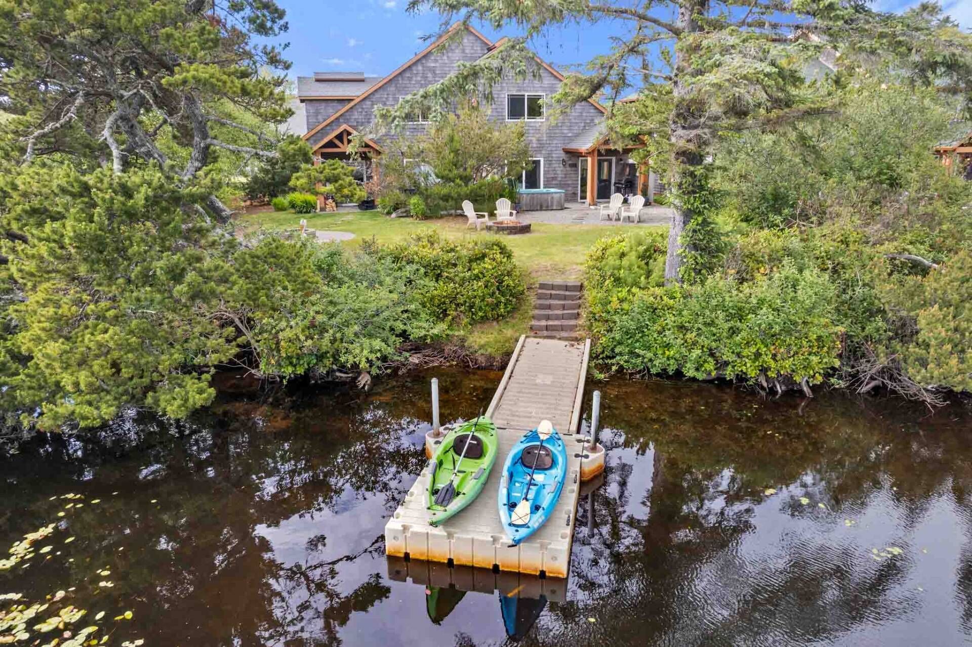 A house with a dock extending into a calm body of water, kayaks docked on the pier, and a well-maintained garden with a pathway leading to the house.
