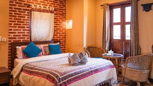 A hotel room with a brick wall, a bed with patterned bedding, two blue pillows, a small round table with a tissue box, and wicker chairs. - Hongsi Hotel (Kathmandu)