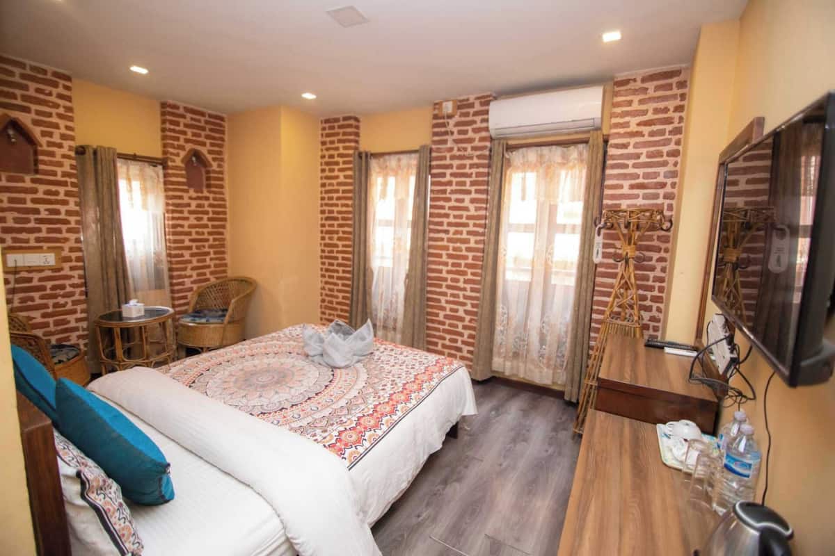 Quarto de hotel com parede de tijolos, cama com colcha estampada, mesa redonda pequena com uma cadeira, televisão fixada na parede e piso de madeira.