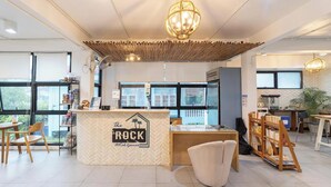 Un interior moderno con una recepción que dice "The ROCK @Koh Gamus", ventanas amplias y un espacio de estar informal con una estantería.