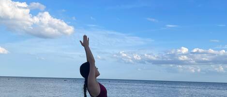Una persona realizando una postura de yoga en la playa, con un cielo despejado y un mar tranquilo al fondo.