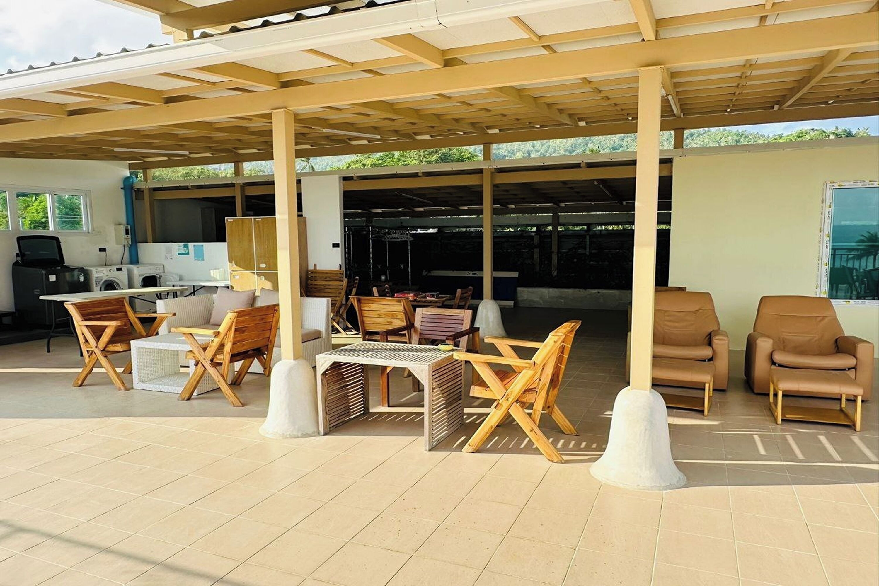 An outdoor covered patio area with wooden furniture, a washing station, and a view of the surrounding greenery.