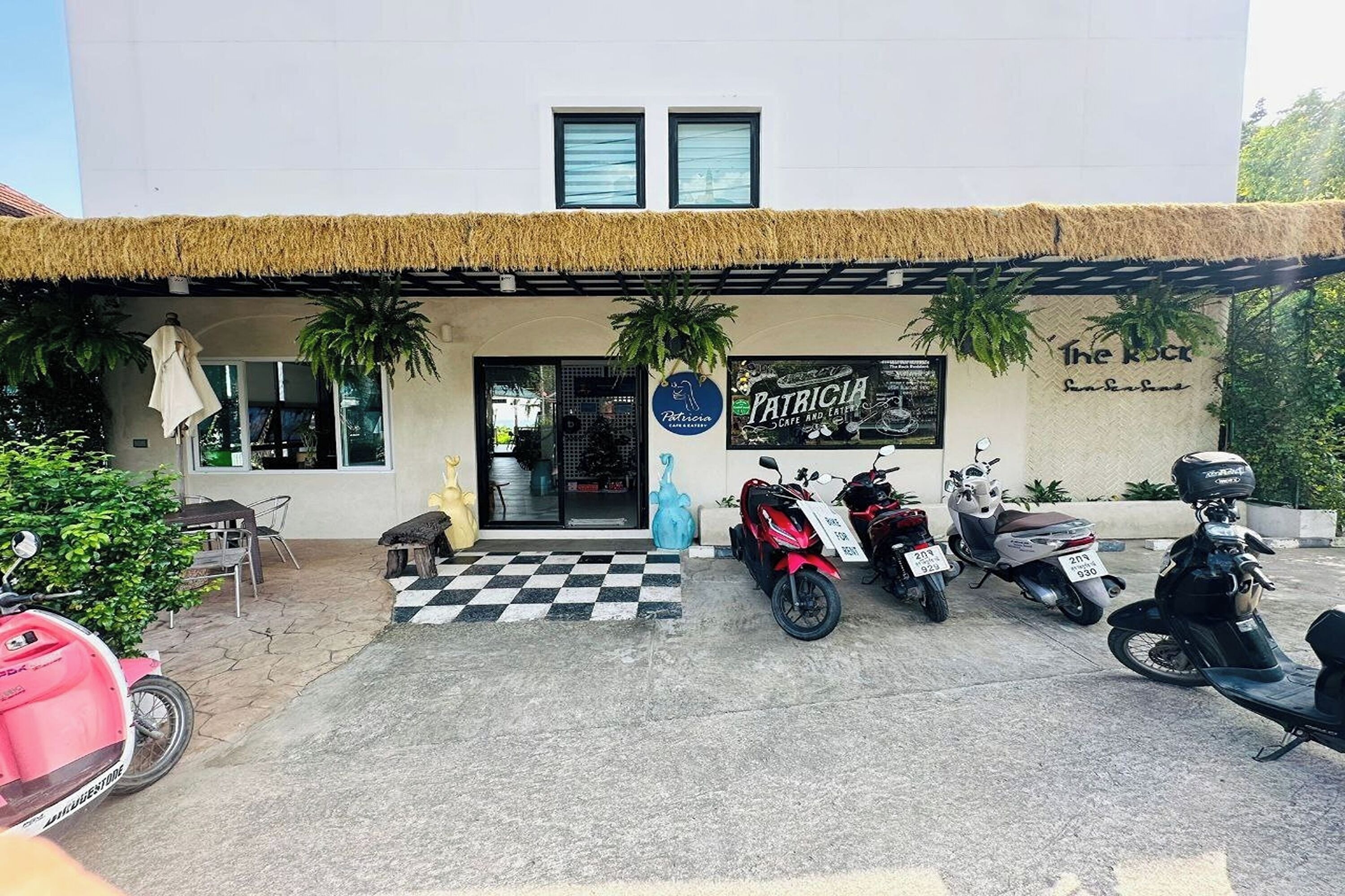 A building with a thatched roof entrance, a sign for 'Patricia Cafe and Eatery', and several parked scooters.