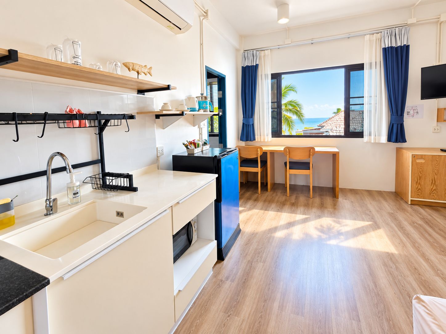 A compact kitchen with a sink, a microwave, and a small refrigerator. There is a dining area with a table and chairs, and a window with curtains.