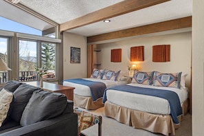 A modern hotel room with two beds, a sofa, a dining area, and large windows offering a mountain view.