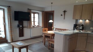 Cocina compacta con armarios de madera, una mesa de comedor y un televisor montado en la pared.