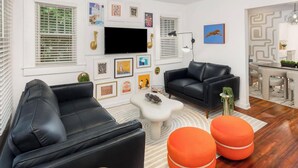 Ein modernes Wohnzimmer mit einer schwarzen Ledersofa, einem weißen Couchtisch und orangefarbenen Hocker. An der Wand ist ein Flachbildfernseher angebracht, und es gibt einen Essbereich mit einem weißen Tisch und Stühlen.