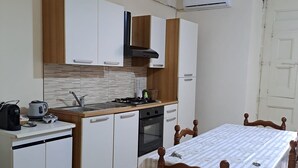 Eine kompakte Küche mit weißen und hölzernen Schränken, einem Gasherd und einem kleinen Backofen. Es ist ein Esstisch mit einem gemusterten Tischläufer und Holzstühlen abgebildet.
