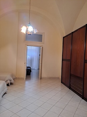 A room with a tiled floor, a bed, a wardrobe, and a door leading to another room.