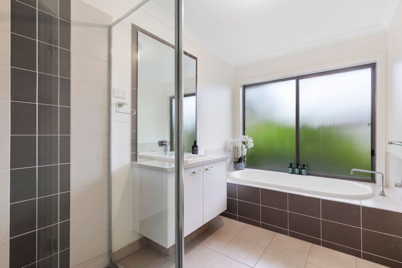 A modern bathroom with a glass shower enclosure, a large window with frosted glass, and a white sink with a mirror.