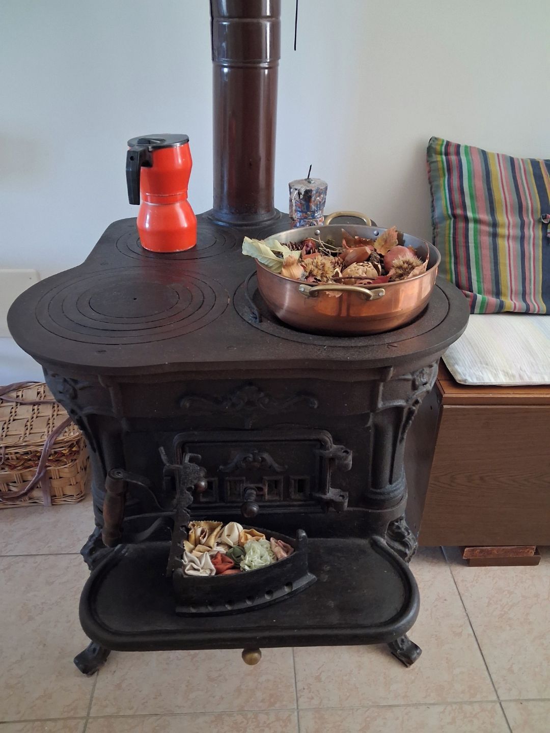 A traditional wood-burning stove with a red pot on top, a copper pot on the stove, and a basket of firewood inside.