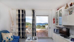 Een woonkamer met een bank, een bijzettafeltje en een televisie. Er is een balkon met uitzicht op de zee en heuvels.