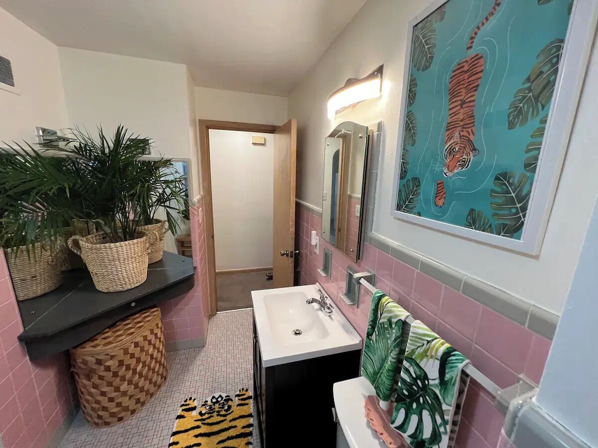 Une salle de bain avec un lavabo blanc, un miroir et un tableau décoratif représentant un tigre accroché au mur. On y trouve des plantes en pot sur une étagère et un porte-serviettes avec une serviette verte.