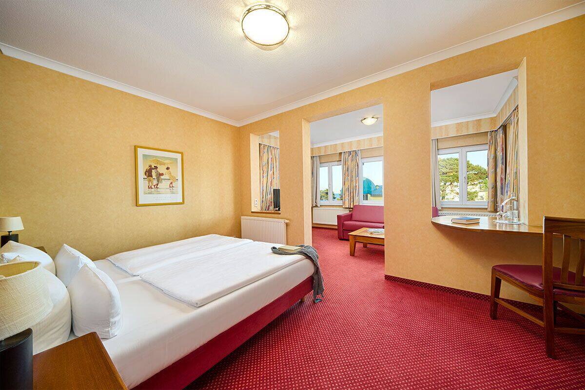 A hotel room with a bed, a desk, a chair, a red carpet, and a window with curtains.