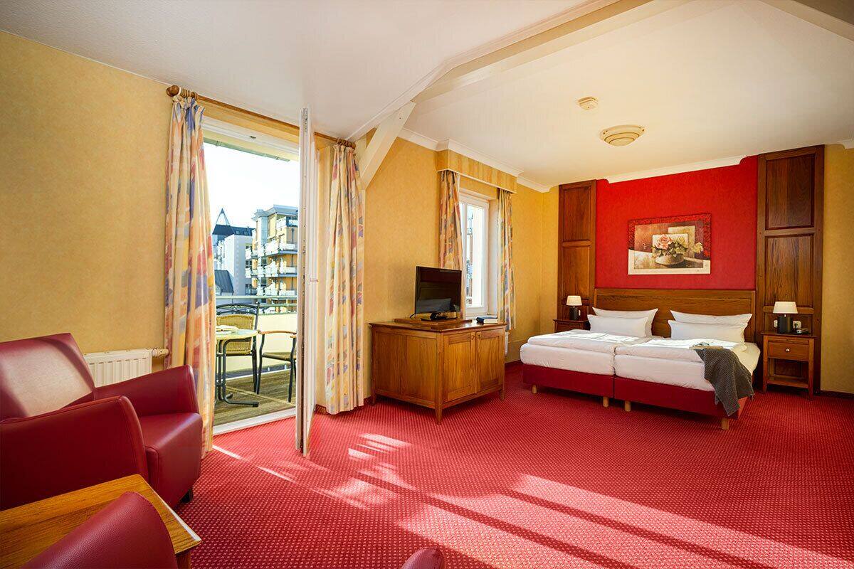 A hotel room with a red carpet, a bed, a desk with a television, a chair, and a window with curtains.