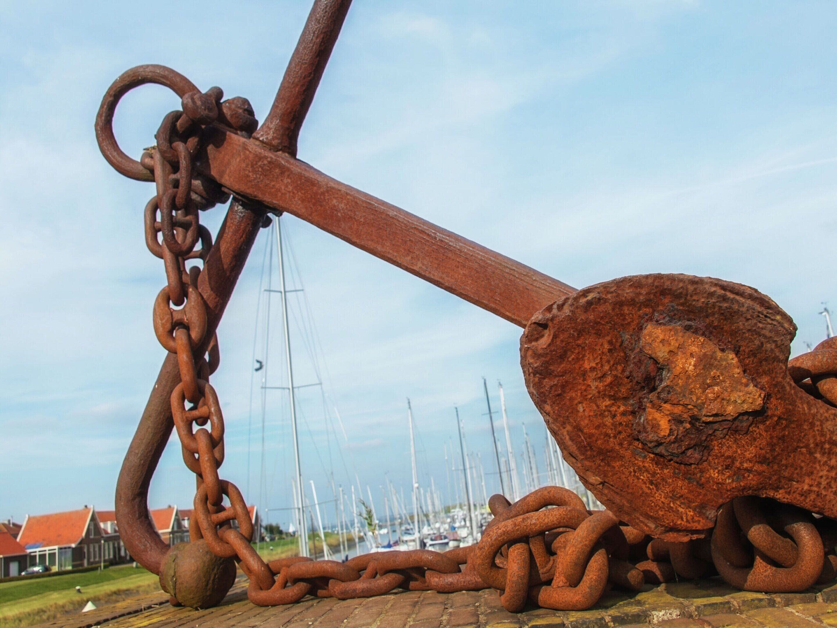 A large rusty anchor with a chain, set against a backdrop of a marina with boats and houses.