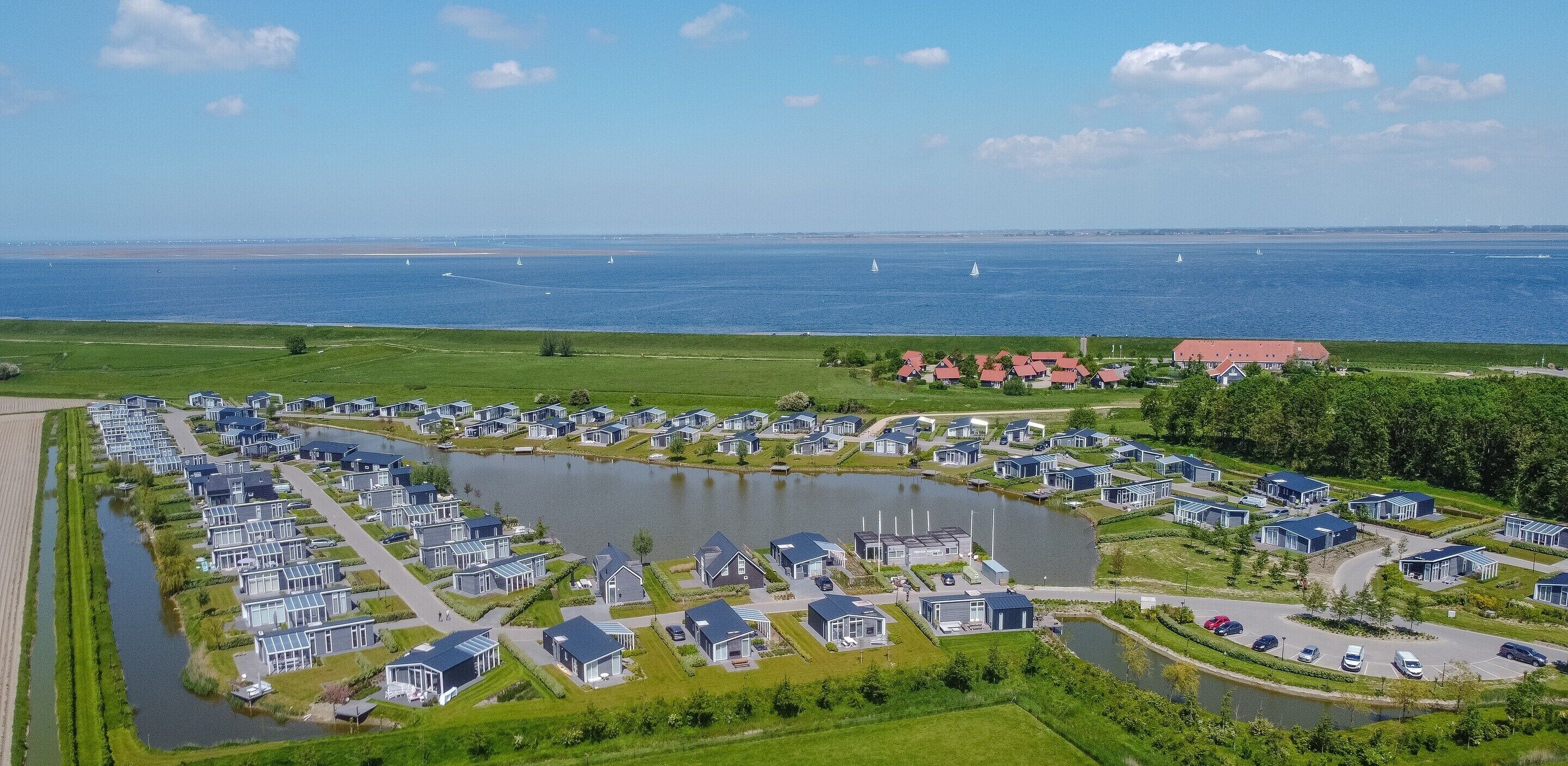 A residential area with modern houses, a central pond, and a view of the sea with sailboats.