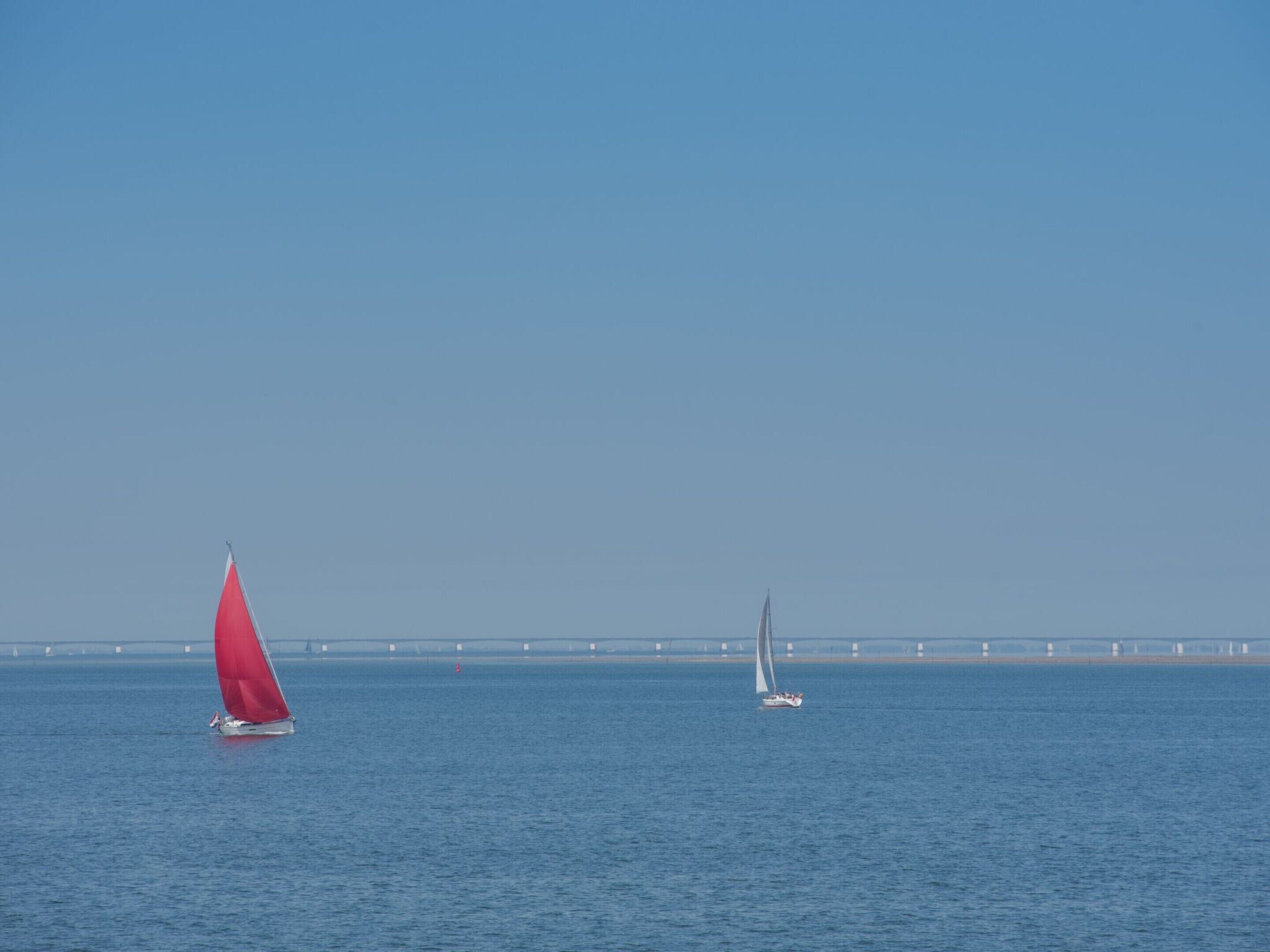 Un voilier à la voile rouge sur une eau calme, un pont en arrière-plan et un ciel dégagé.
